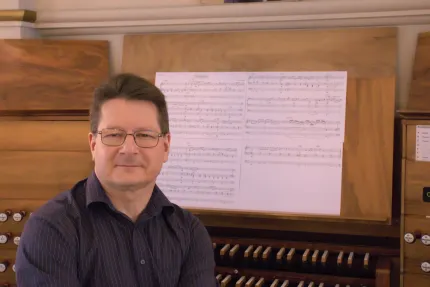 En man med glasögon sitter vid en orgel med notblad uppställt på notstället i en träinredning..
