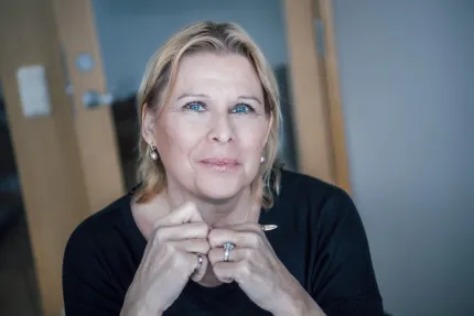Närbild av en blond kvinna som sitter inomhus med händerna knäppta vid hakan och ser in i kameran..