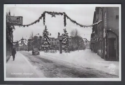Svartvit vy över snötäckt stadsgata med bil, gångtrafik och granar; julgirlanger hänger över vägen..