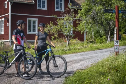 En man och en kvinna i cykelkläder och hjälmar står på en grusväg framför ett rött hus och håller i varsin cykel.
