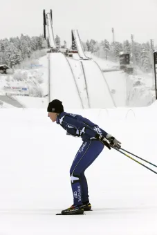 Längdskidåkare som stakar, hoppbackar i bakgrunden.