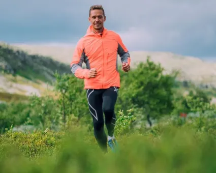 Man springandes i fjällmiljö med orange vindjacka och svarta byxor.