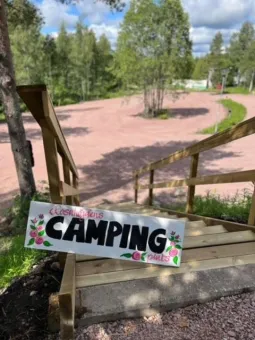 En skylt med texten Rosahöjdens camping plats placerad i en trä trappa, i bakgrunden en grusplan ogiven av träd.