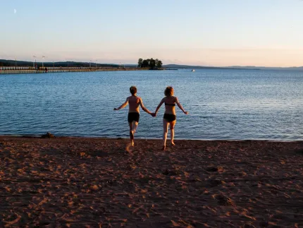 En man och en kvinna som springer hand i hand på en sandstrand på väg ner i vattnet, Långbryggan i bakgrunden.