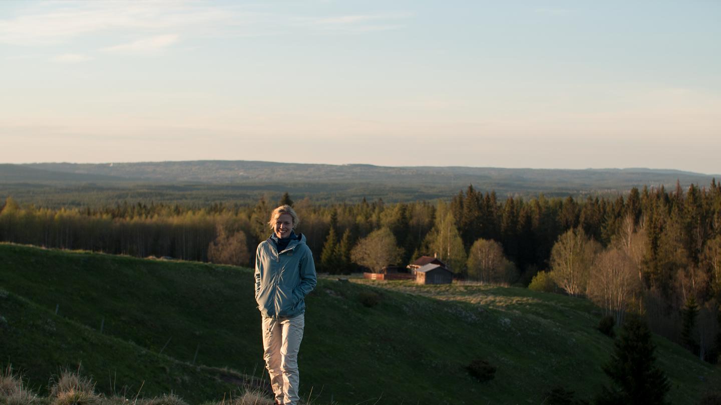 En kvinna står i ett öppet landskap med utsikt över skog och berg.