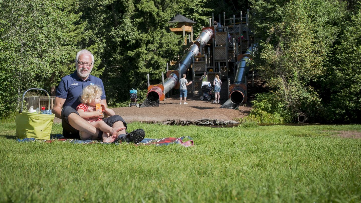 En Morfar som sitter med sitt barnbarn och fikar i en lekpark.