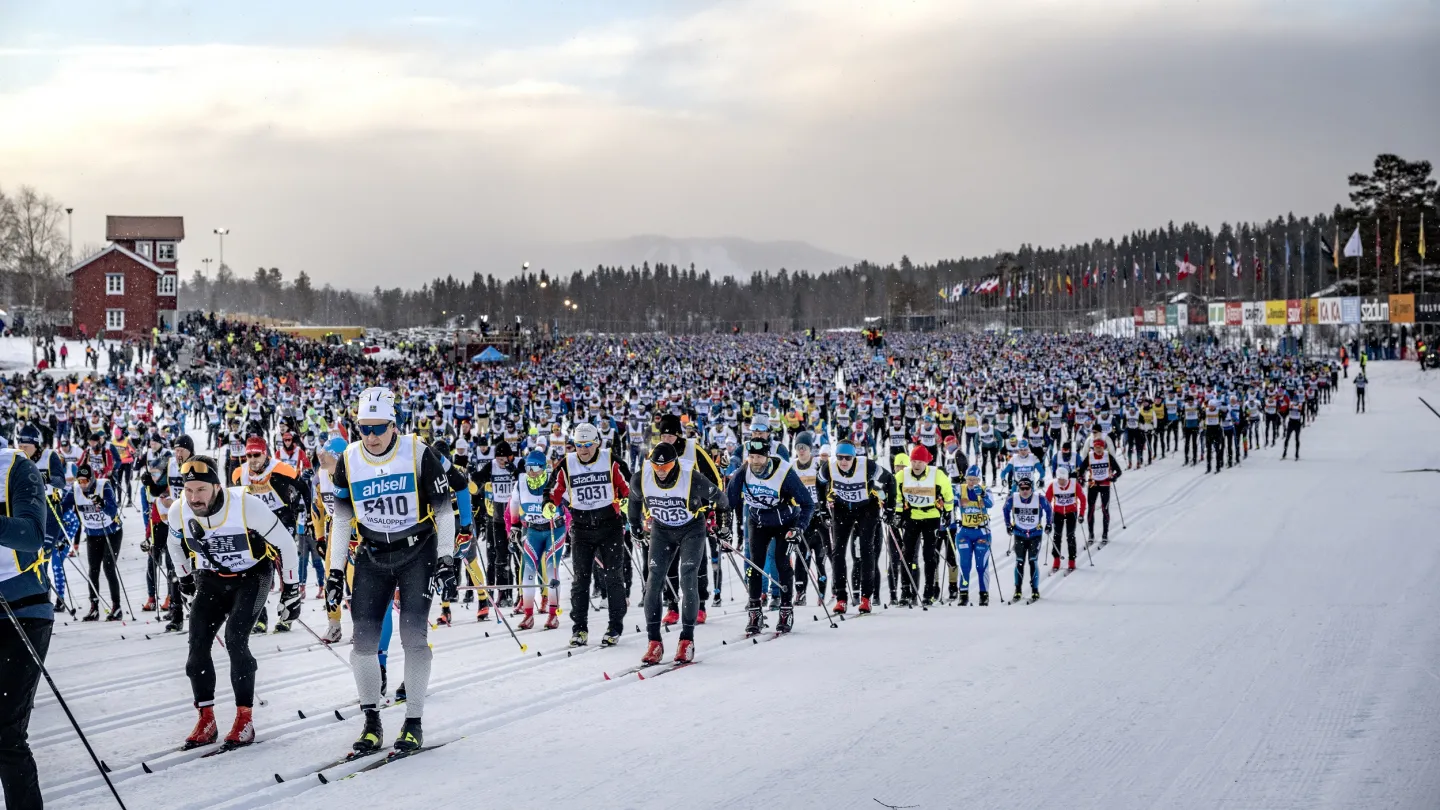 A large crowd of cross-country skiers at a mass start on a snowy course, with flags and a red building nearby.