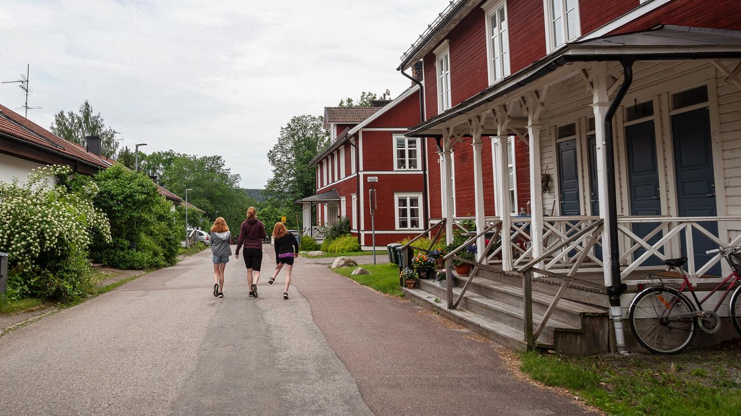 Två barn och en vuxen går längs en gata bredvid ett rött hus.