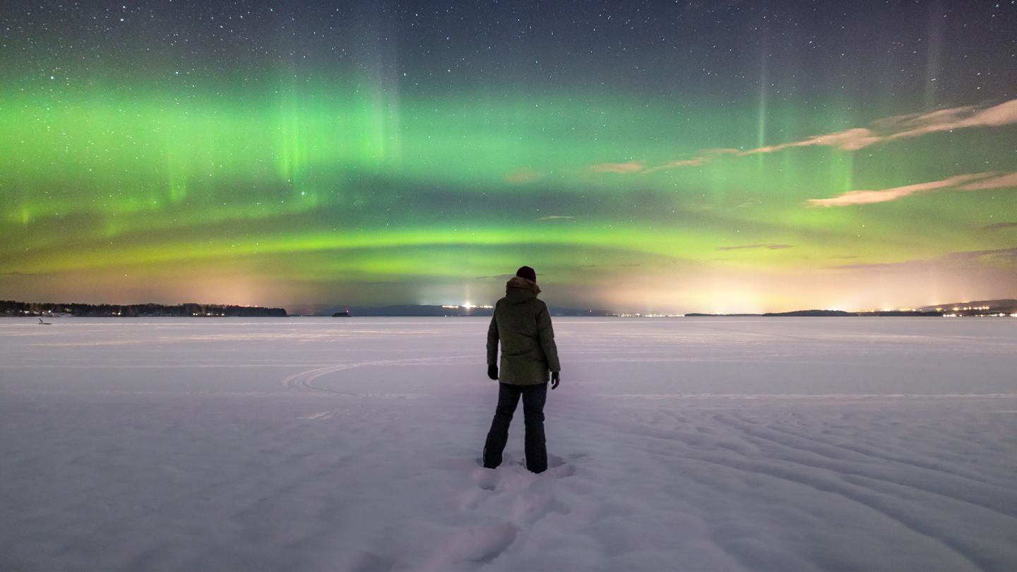 En man står på en snötäckt sjö, över himlen sprakar ett grönt norrsken.