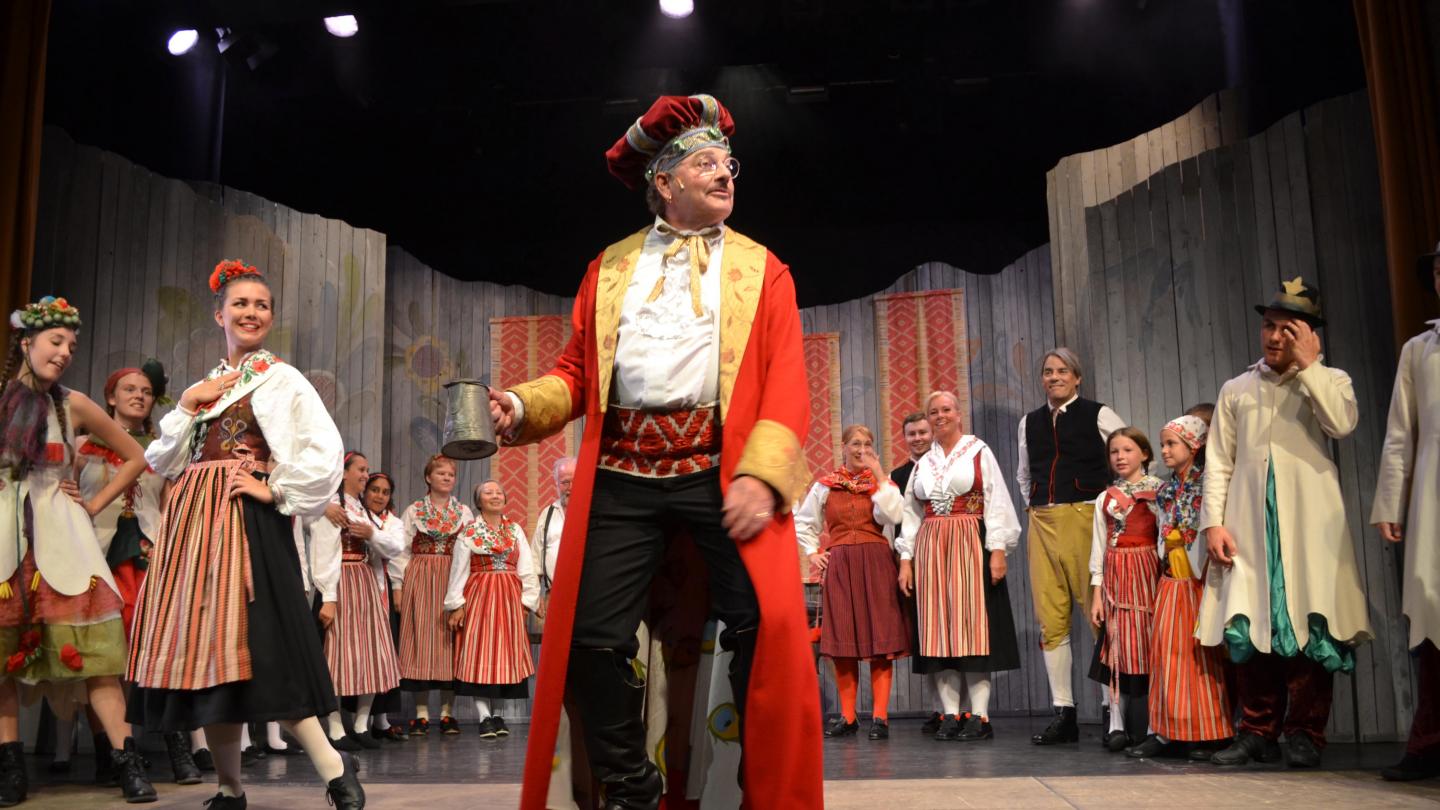 En teaterscen med människor i folkdräkt, i mitten en man i röd kappa och hatt.
