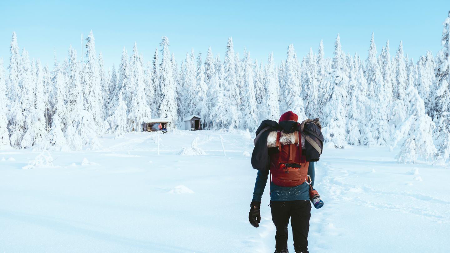 En person med ryggsäck går mot en stuga i snötäckt skog.